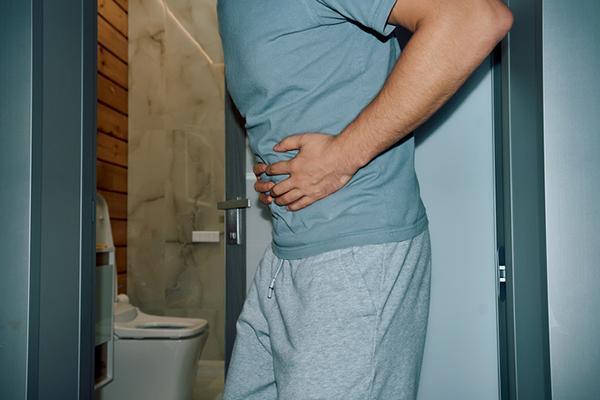 Cropped image of the midsection of a man gripping his stomach with both hands as he stands in front of a bathroom.