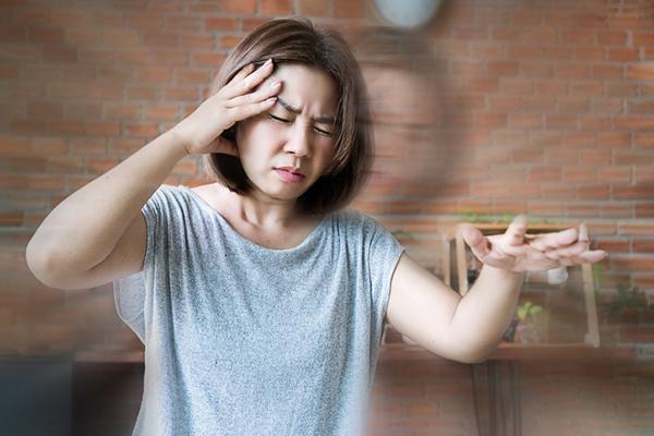 A woman experiencing dizziness holds one arm out in front of her and her other hand to her head. her eyes are closed and there is a double-image effect to the photo.