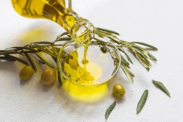 High-angle view of olive oil being poured from a glass carafe into a glass bowl, with a fruit-bearing olive branch next to it.