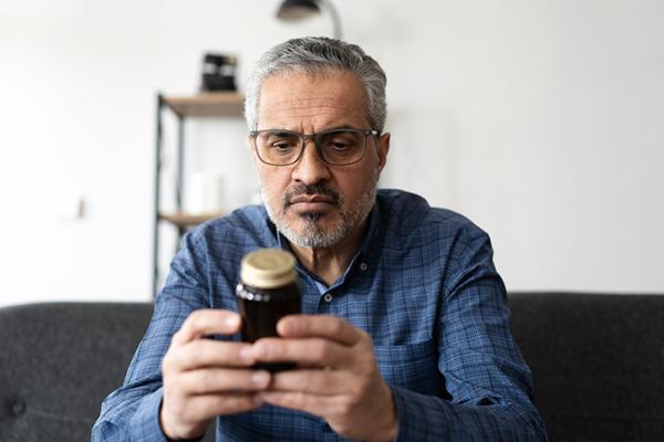 A mature man sits on his couch, holding a brown bottle containing vitamins and reading the label.