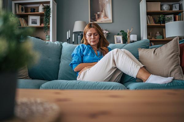 A woman experiencing intense abdominal pain while sitting on the sofa at home, holding her belly with both hands and showing signs of discomfort.