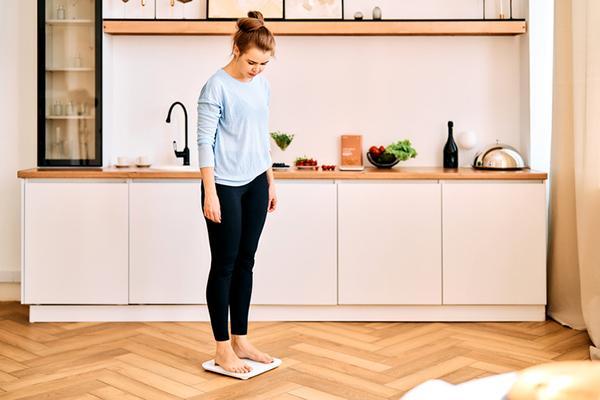 A woman stands barefoot on a digital scale in her kitchen, looking down at the display.