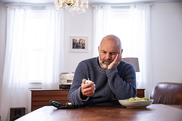 A middle-age man sits at a dining table in his home, looking at a blood sugar monitor in his hand with a salad on the table next to him.
