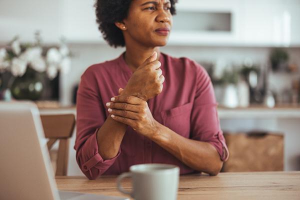 A middle-age woman sitting at a table in the kitchen holds her wrist and looks like she is in pain.