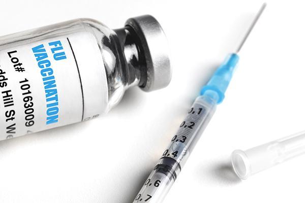 A vial of flu vaccine on a white surface next to a syringe with the cap removed.