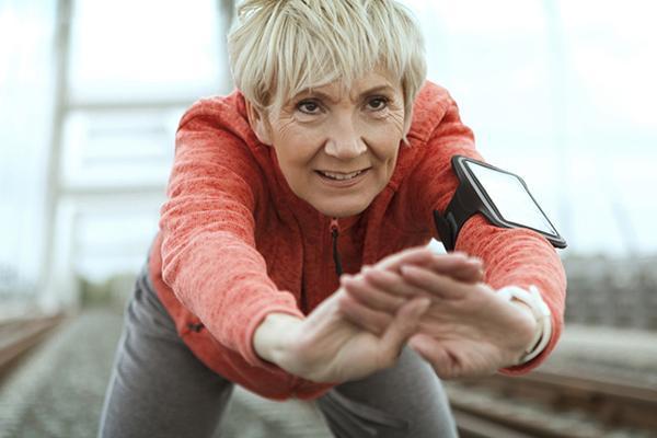 Close-up of a mature woman paused during a run to lean forward and stretch; behind her, out of focus, appears to be a bridge.