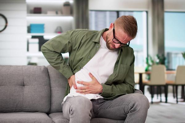 A man sitting on his couch at home, experiencing intense pain and holding his right side with both hands.