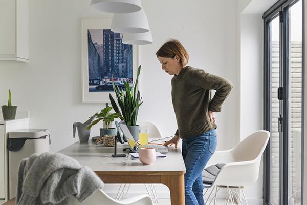 A middle-age woman stands up from her desk while working from home, wincing from back pain.