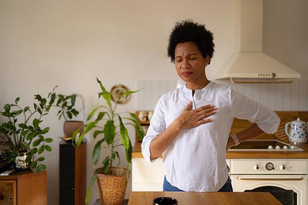 A woman stands in her kitchen, experiencing chest pain with one hand on her upper chest and a look of discomfort on her face.