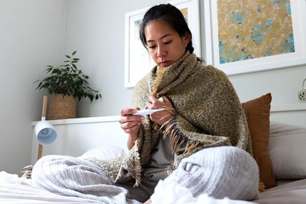 A woman sitting on a bed bundled in several layers of clothing looks at a digital oral thermometer.