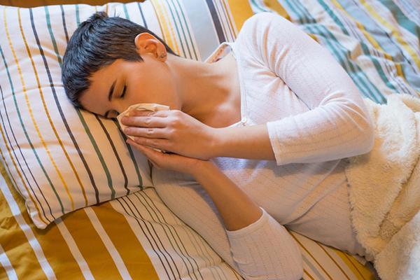A young woman in bed with a cold, blowing her nose while lying on her side.