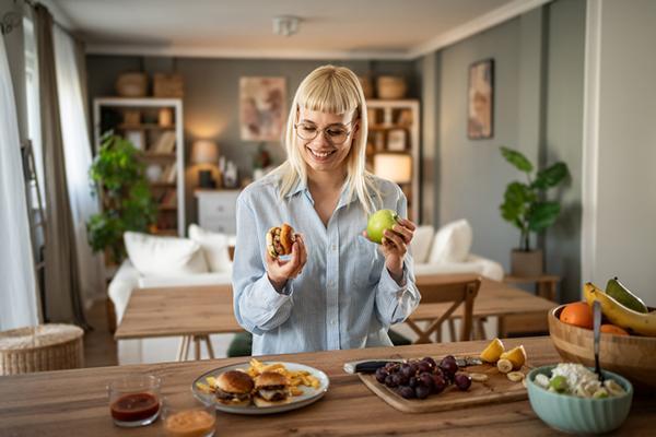 A smiling woman stands at a buffet spread on a counter, holding a burger in one hand and an apple in the other.