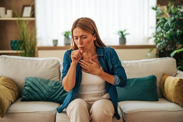 A middle-age woman sits on a couch, coughing; one hand is on her upper chest and the other is in front of her mouth.