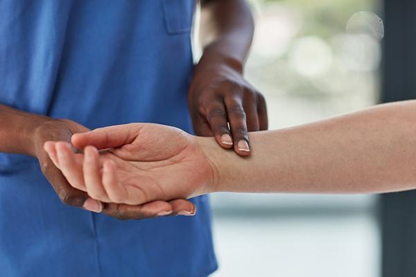 Cropped image shows the torso of a medical professional in a blue scrub top taking the pulse at the wrist of a patient.
