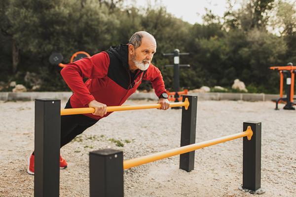 A senior man performs resistance exercises using uneven parallel bars in an outdoor setting.
