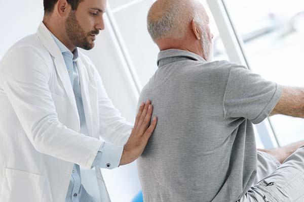 A doctor examining a senior man encourages him as he tries to touch his toes while sitting with his legs extended out in front of him.