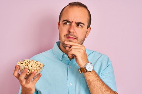 A man holds a dish of pistachios in one hand and the other to his chin, looking thoughtful.