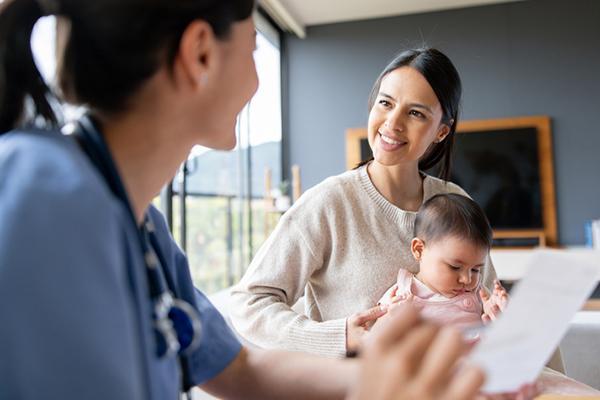 A smiling mother holds her baby in her lap while talking with a pediatrician during what appears to be a home visit.