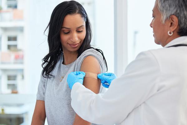 A woman looks down at her arm as she watches a health care provider place a bandage over the site of a vaccination.