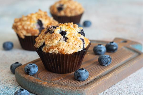 A blueberry muffin on a wood cutting board with some fresh blueberries scattered around it, and two more muffins in the background, out of focus.
