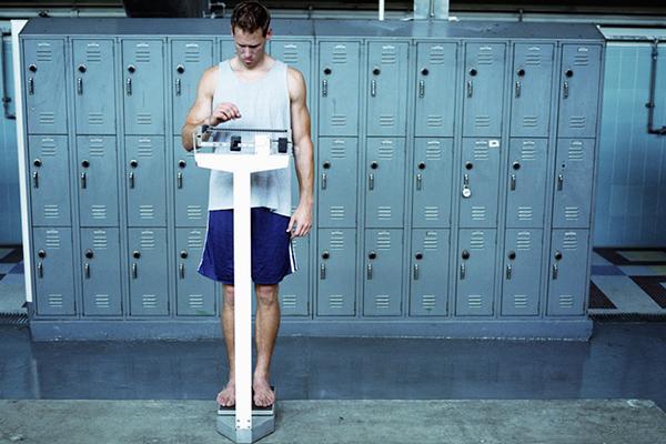 A young man in a tank top and athletic shorts weighs himself using an upright scale in a locker room.