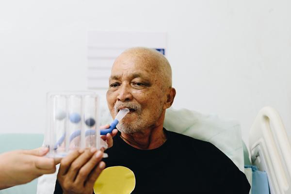 A senior man breathes into a tube for a lung function test as a hand from an unseen person holds the device steady.