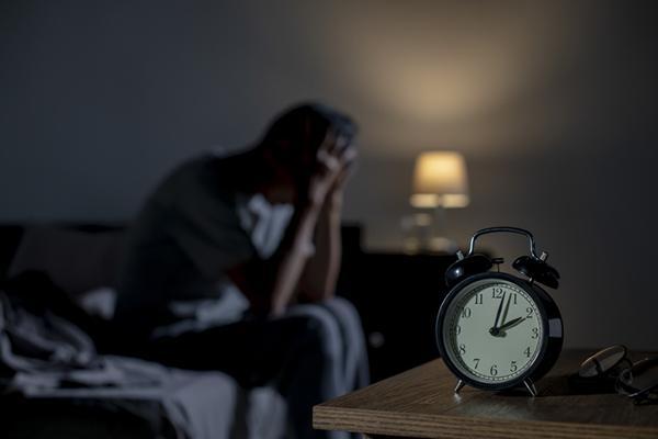 An alarm clock on a table reads 2 AM; in the background, out of focus, a man sits on a bed resting his head in his hands.