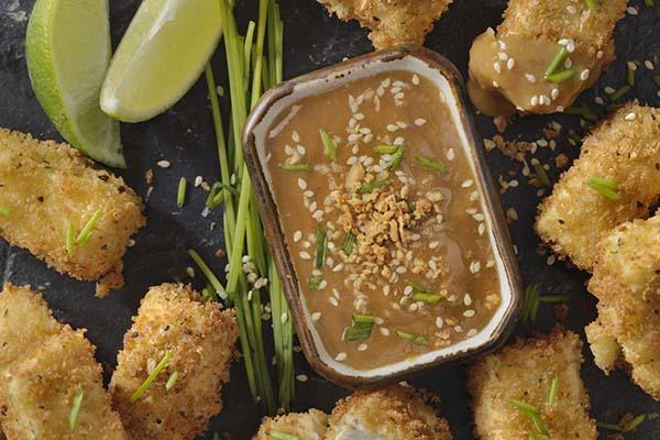 Breaded tofu bites viewed from above, with a ginger peanut dipping sauce in a dish in the middle and wedges of lime top left.