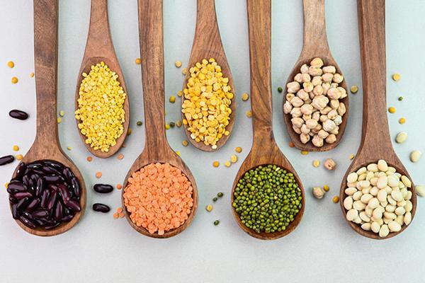 A row of seven wood spoons holding various types of legumes including black beans, lentils, and peas.