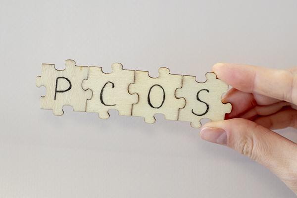 A hand holds four connected puzzle pieces with the letters P C O S written on them, representing polycystic ovary syndrome.