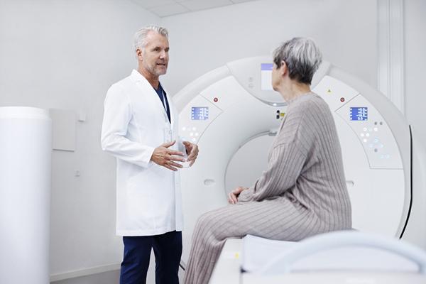 A doctor talks with a senior woman who is sitting on the table of a scanning machine, about to undergo a screening.
