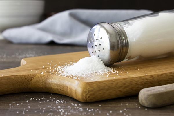 Close-up of a salt shaker tipped on its side on a wood cutting board, with salt spilled out.