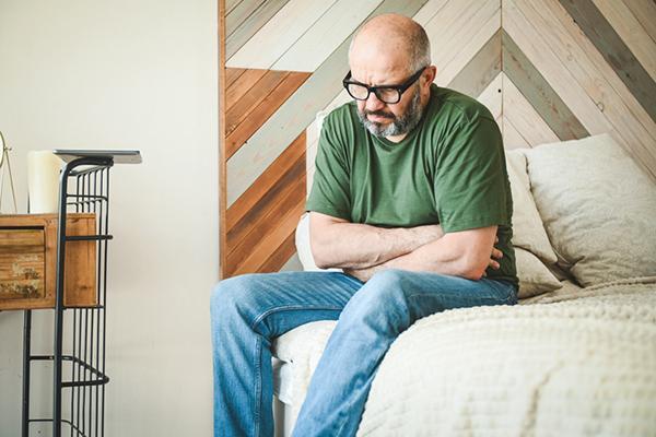 A middle-age man sits on a bed with his arms across his midsection, in obvious discomfort.