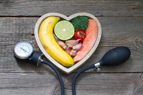 Healthy food choices arranged in a heart-shaped dish, with a blood pressure gauge positioned under it on a wood table.