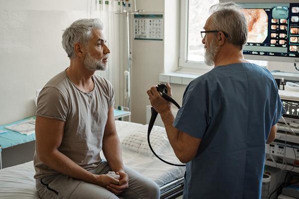 A doctor explains colonoscopy results to a patient using monitor images on a screen.