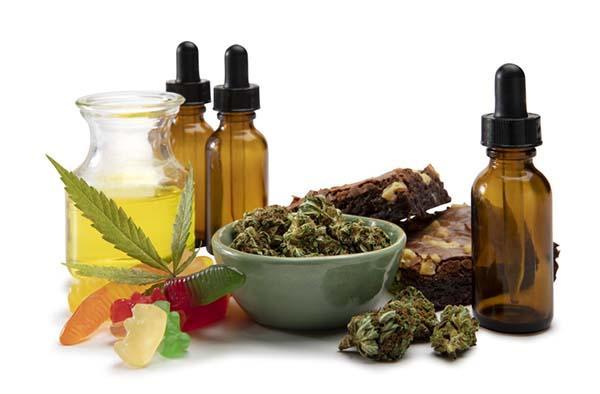 A bowl containing cannabis buds is surrounded by several glass bottles with eye dropper tops and a small carafe containiing oil; gummies; and brownies.