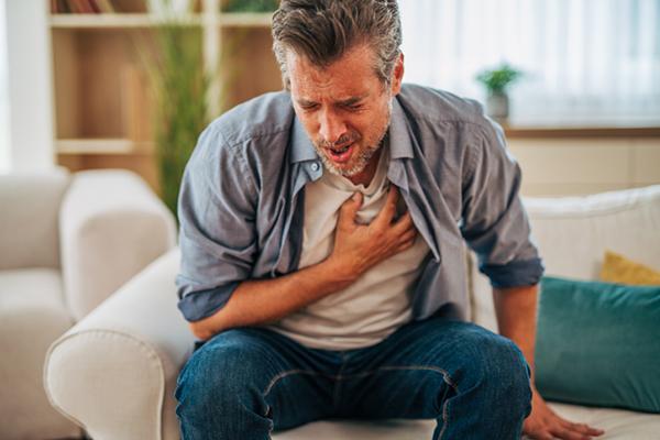 A mature man leans forwars and grips his chest, experiencing pain as he sits on a sofa at home.