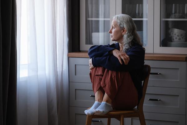 A mature woman sits on a chair in a dimly lit room with her knees pulled up to her body and her feet resting on the chair seat, looking toward a curtained window.
