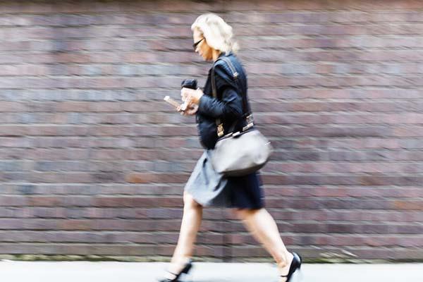 Motion-blurred image shows a woman wearing business attire walking quickly in front of a brick wall while holding her phone in one hand and a coffee cup in the other.