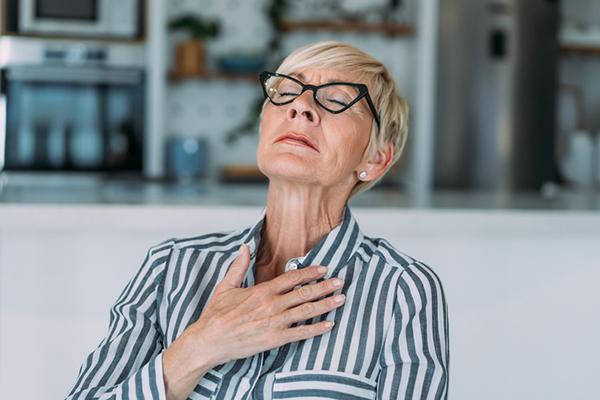 A mature woman holds a hand to her chest and tilts her head back as she experiences chest pain.