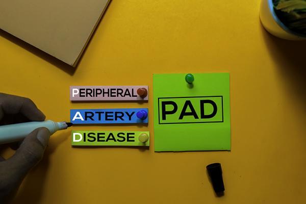 Strips of paper with the words peripheral artery disease, held to an orange surface with push pins; to their right is a green post-it with the letters P A D, also held up with a push pin.
