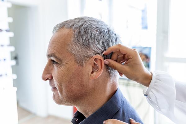Side view of a mature man being fitted for a hearing aid; the device is an over-the-ear type with a small element on a wire in his ear.