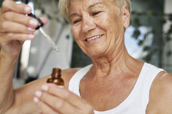 A senior woman holds a glass bottle of facial serum in one hand and the dropper lid in the other.