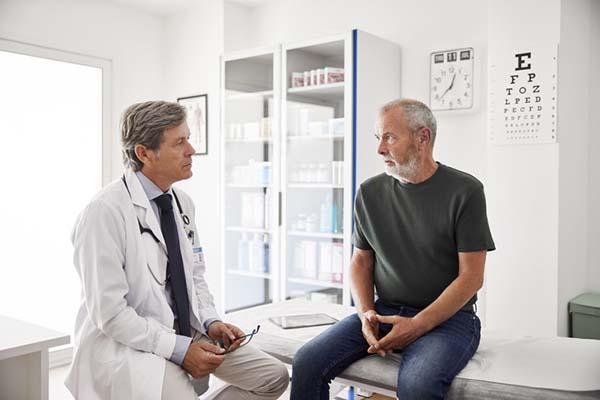 A senior man talks with his doctor while sitting on the table in an examination room.