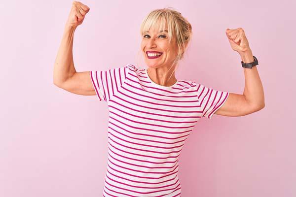 A middle age woman wearing a striped shirt smiles broadly and flexes her arms.