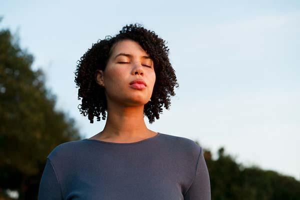 A woman stands outdoors with her eyes closed, concentrating on taking in the sounds of nature.