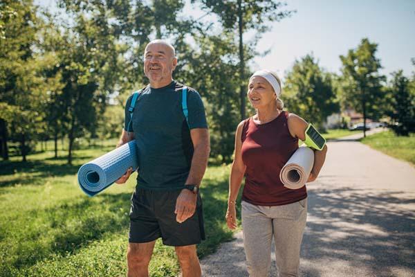 A mature couple walking in a park and carrying exercise mats, heading to an outdoor yoga class.