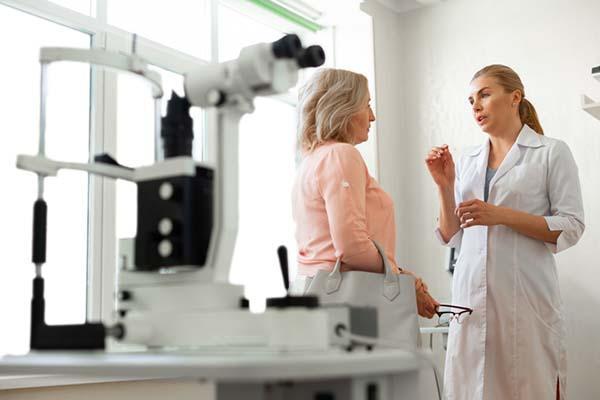 An eye doctor speaks with a middle-age patient in an exam room with equipment visible in the foreground.