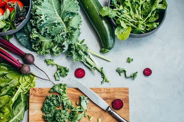 Overhead view of fresh leafy greens, cucumber, beets, and other vegetables arranged next to a cutting board and knife.