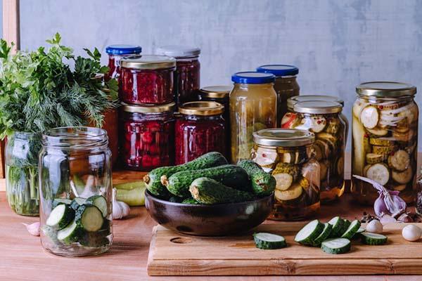 A bowl of fresh cucumbers rests on a cutting board where another cucumber has been sliced, next to jars filled with pickles and beets and an open jar with more cucumber slices.
